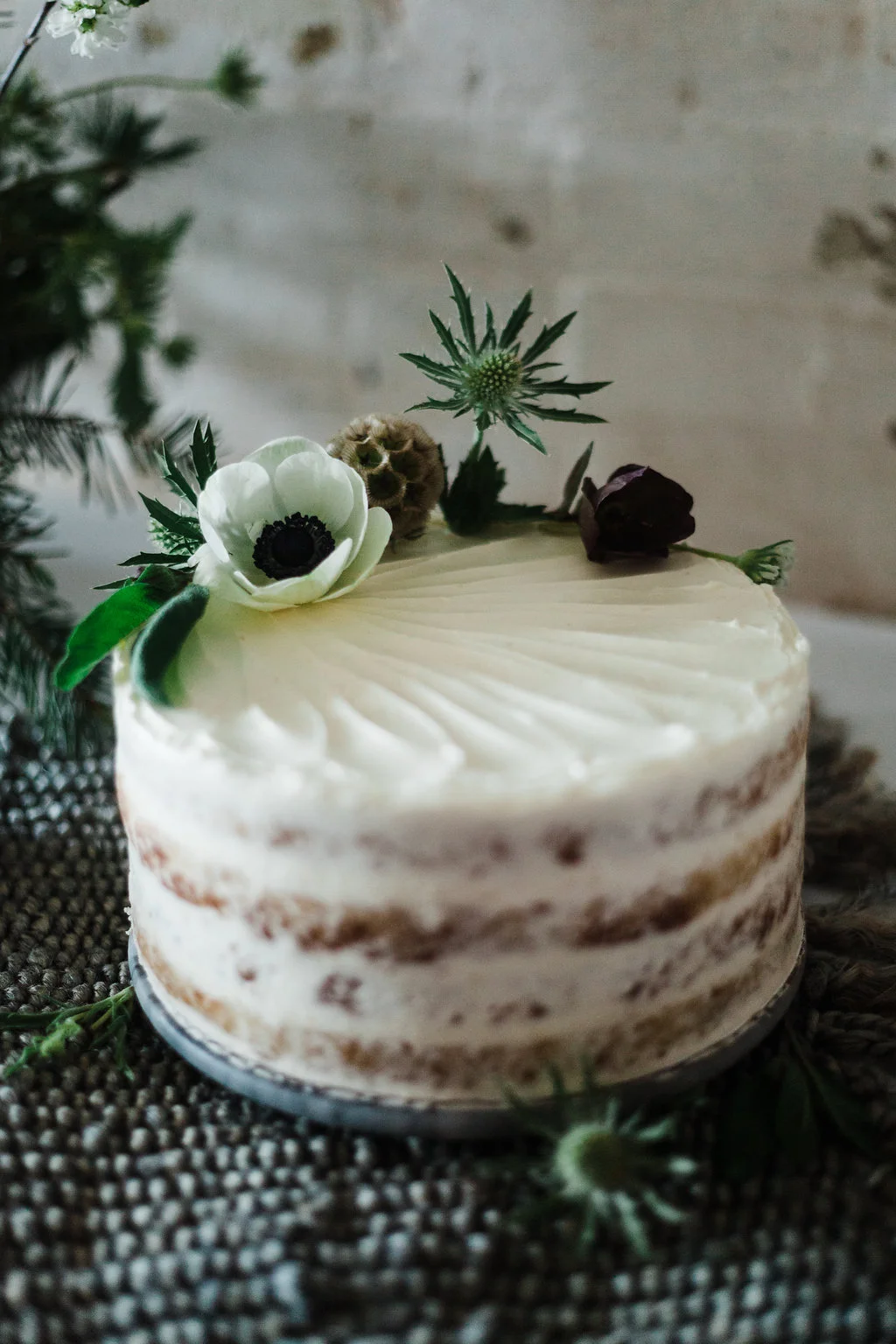 Small round semi-naked cake topped with white anemone and dark flower on textured dark cloth