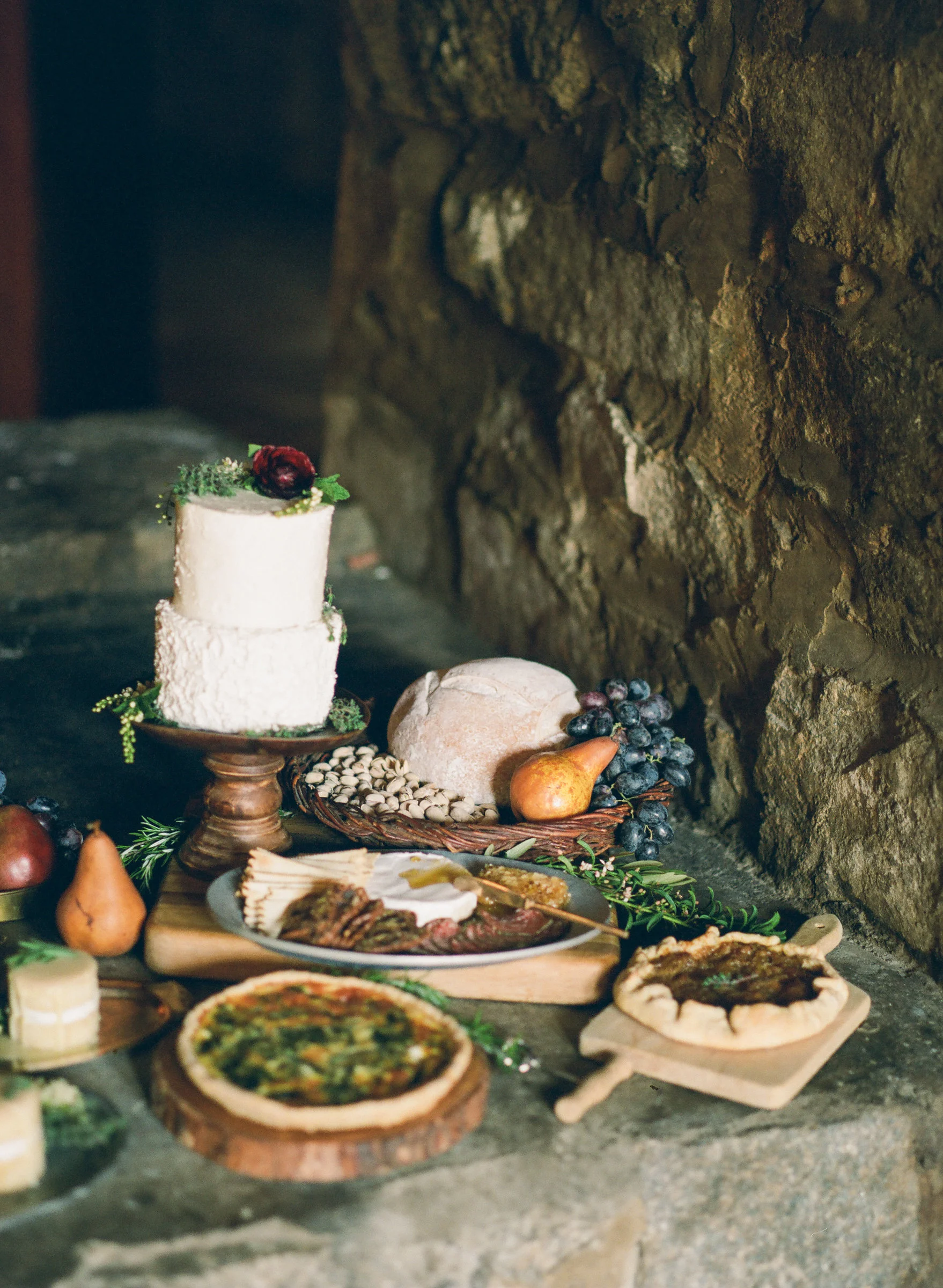 Rustic food spread including a tiered white cake, bread, nuts, fruit, cured meats, and tarts arranged on stone steps, warm earthy tones