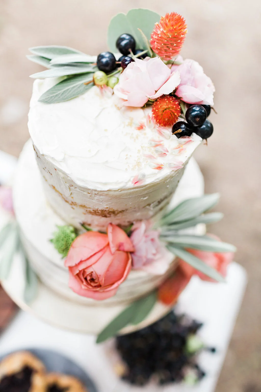 Overhead view of a tiered white frosted cake decorated with roses, berries, and greenery, soft neutral tones with pink accents